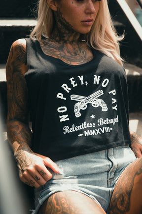 A tattooed person with long blonde hair sits on outdoor steps, wearing the Relentless Betrayal No Prey, No Pay Womens Crop Tank featuring crossed pistols and paired with relaxed-fit denim shorts.