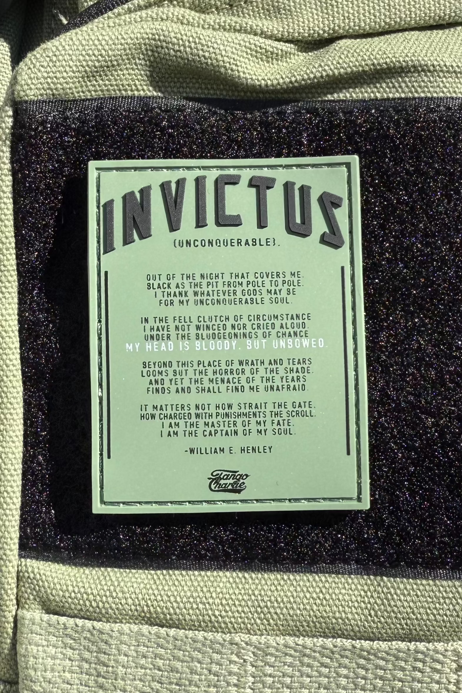 The Invictus PVC Patch by Tango Charlie Apparel features William E. Henley’s poem in black text on green, set against textured light beige fabric with a dark glittery strip—ideal for fans of inspirational gear and messages of strength.