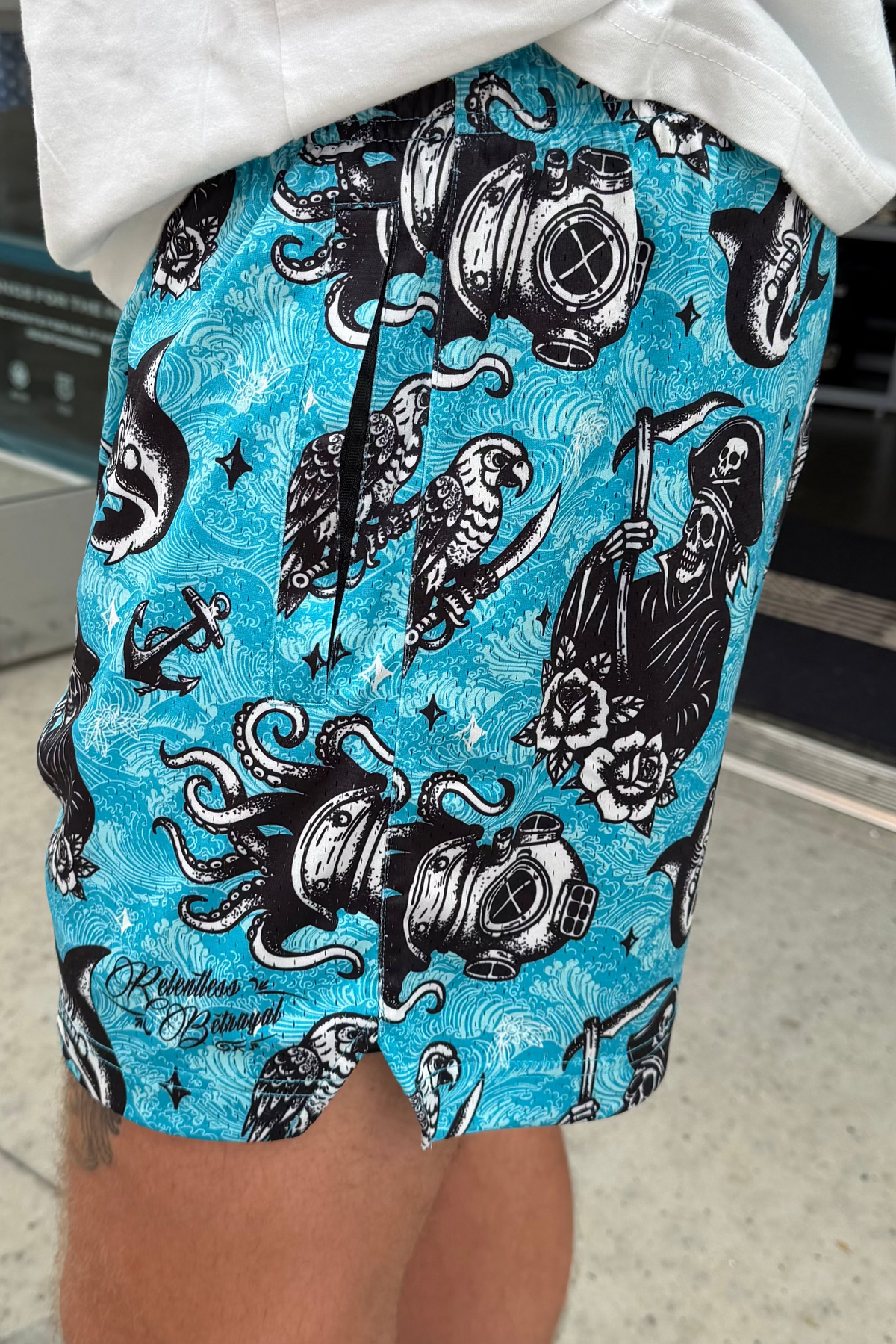 A close-up of someone outdoors in Relentless Betrayal's Premium Mesh Shorts | 5.5"- Davy Jones, featuring bright blue fabric with black and white octopuses, diving helmets, skulls, parrots, anchors, roses, and waves.