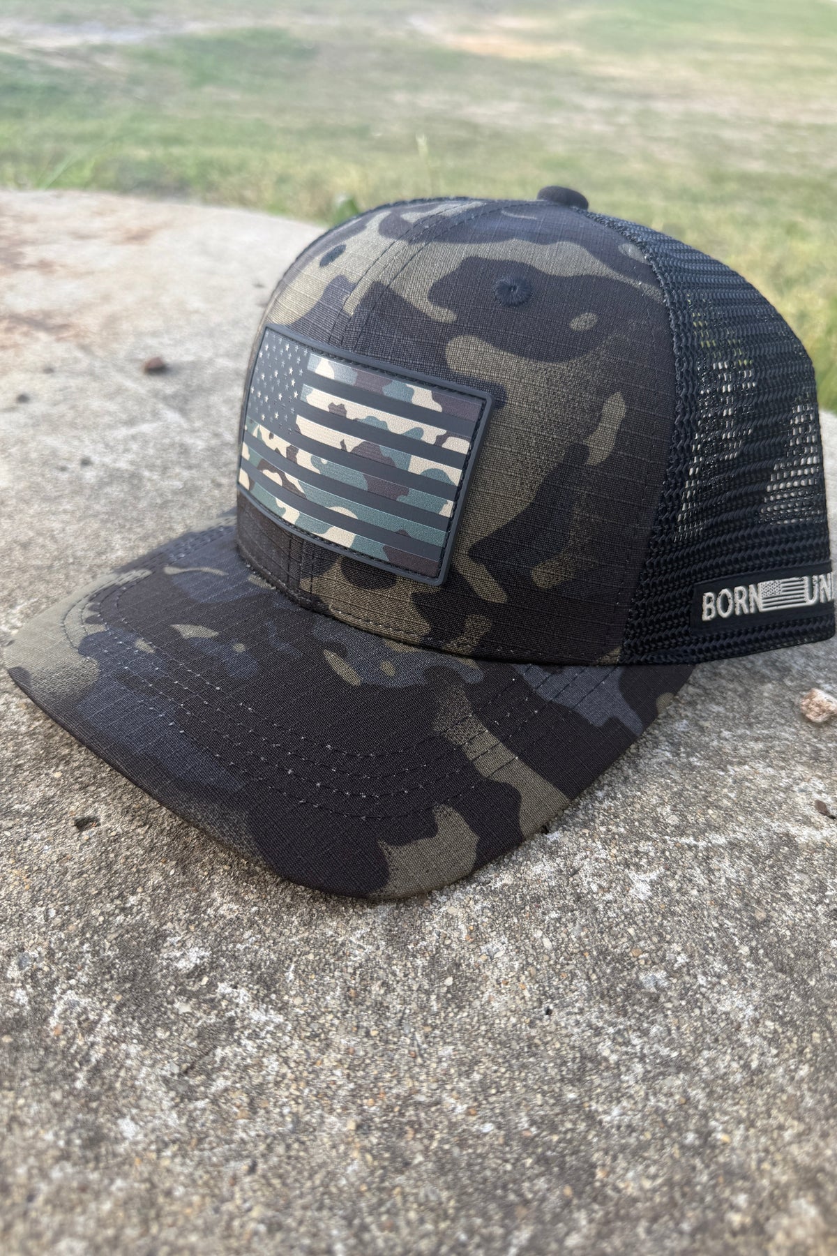 The Born United Black Camo Trucker - Camo Flag by Born United Private Label features a camo-patterned front with a matching camo American flag patch, mesh back, and "BORN" on the side band, perfect for standout patriotic style.