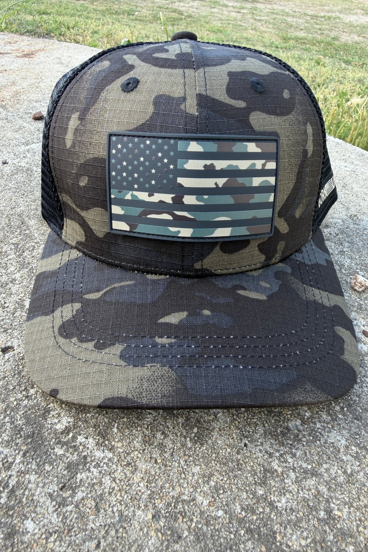 The Born United Private Label "Born United Black Camo Trucker - Camo Flag" snapback features a black camo design with a matching camouflage American flag patch on the front, shown resting outdoors on concrete.