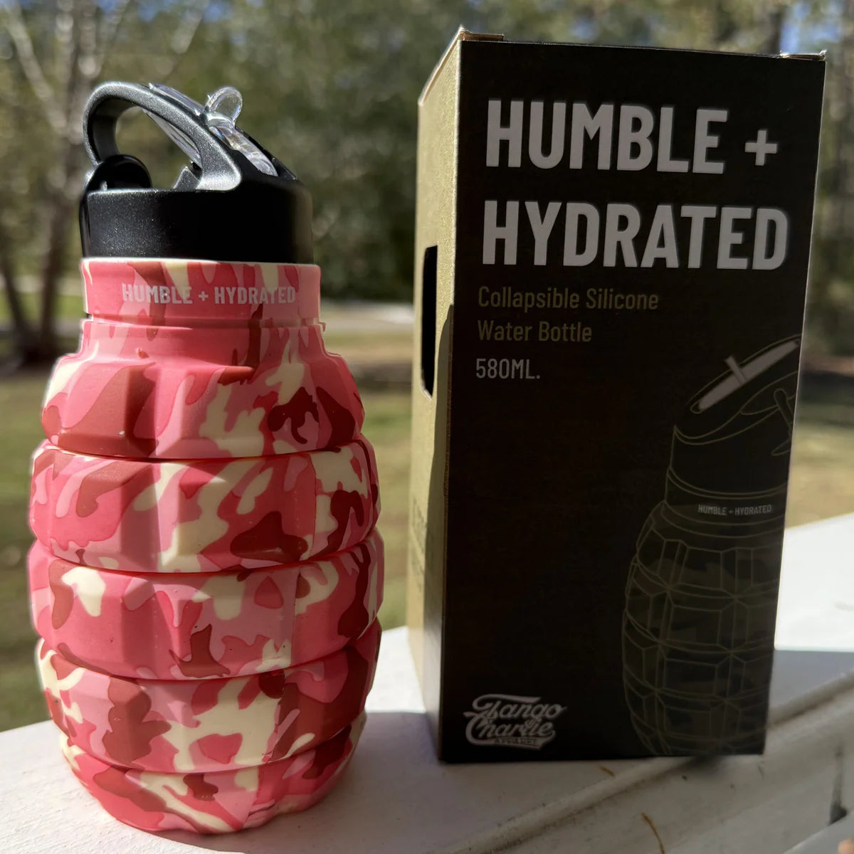 A Pink Camo Humble + Hydrated 20oz Water Bottle by Tango Charlie Apparel, shaped like a grenade and BPA-free, sits next to its box with trees and sunlight in the background.