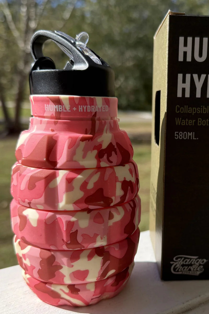 A Pink Camo Humble + Hydrated 20oz Water Bottle by Tango Charlie Apparel, shaped like a grenade and BPA-free, sits next to its box with trees and sunlight in the background.