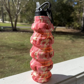 The Pink Camo Humble + Hydrated 20oz Water Bottle by Tango Charlie Apparel, made of BPA-free silicone with a black cap and carabiner, sits on a white railing outdoors against a backdrop of blurred trees and grass.