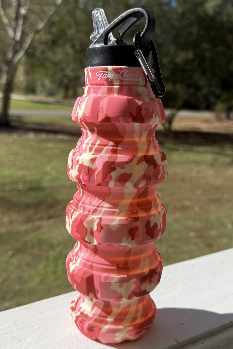 The Pink Camo Humble + Hydrated 20oz Water Bottle by Tango Charlie Apparel, made of BPA-free silicone with a black cap and carabiner, sits on a white railing outdoors against a backdrop of blurred trees and grass.