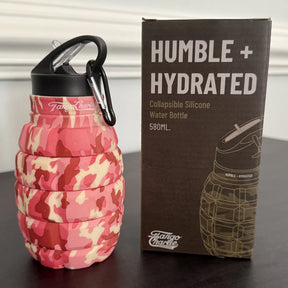 A pink camo Tango Charlie Apparel Humble + Hydrated 20oz collapsible water bottle, shaped like a grenade with a black carabiner clip, stands next to its box. Made from food-grade silicone.