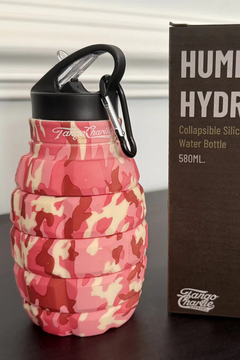 A pink camo Tango Charlie Apparel Humble + Hydrated 20oz collapsible water bottle, shaped like a grenade with a black carabiner clip, stands next to its box. Made from food-grade silicone.