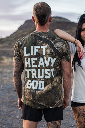 A man stands outdoors facing away, wearing the Tango Charlie Apparel Lift Heavy. Trust God. - Realtree Premium Camo Tee with bold white text. A woman's arm rests on his shoulder, set against rocky terrain and hills.