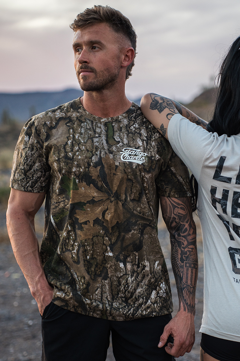 A man wears the "Lift Heavy. Trust God." Realtree Premium Camo Tee by Tango Charlie Apparel, standing outdoors at dusk with mountains behind him. A tattooed woman, mostly out of frame, rests her arm on his shoulder as he looks to the side.