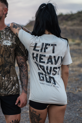 A woman with long dark hair in a ponytail wears the "Lift Heavy. Trust God. - Tee" by Tango Charlie Apparel, standing beside a tattooed man in camo. Smiling outdoors on a rocky path, they embody Christian fitness inspired by Proverbs 3:5-6.