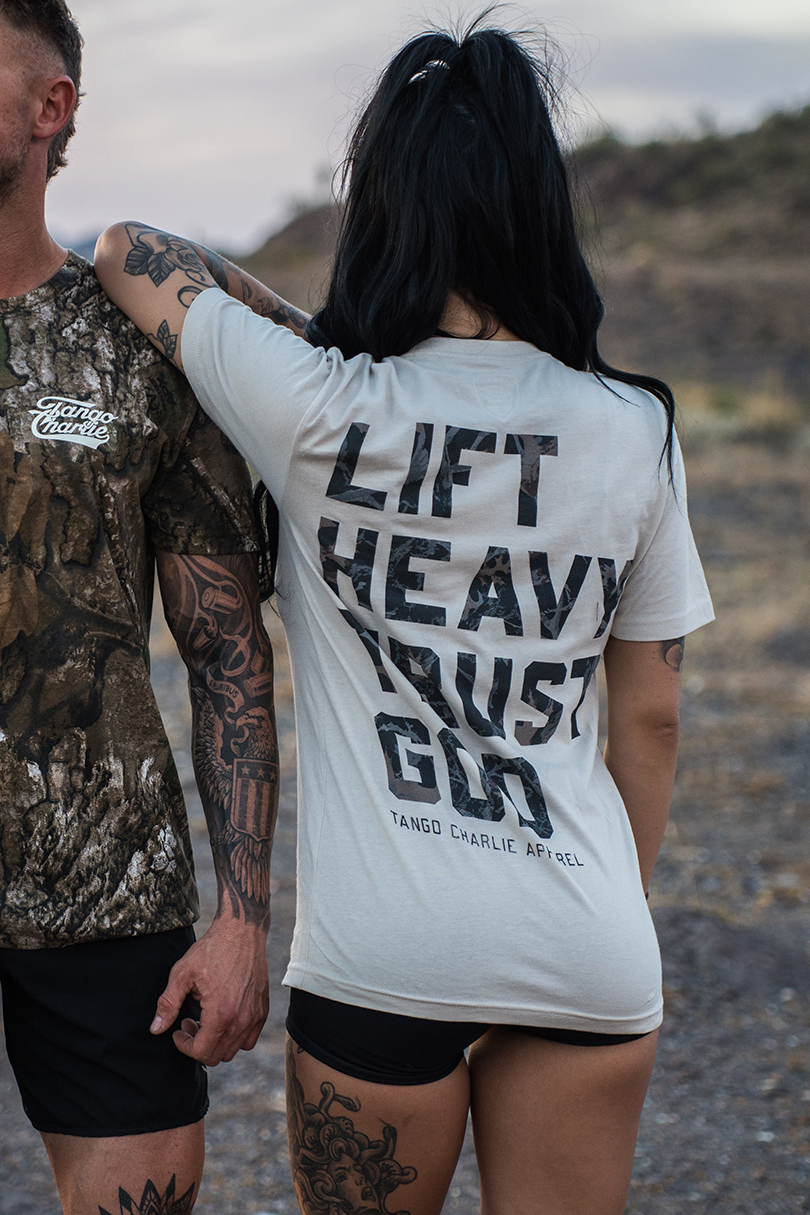 A woman with long dark hair in a ponytail wears the "Lift Heavy. Trust God. - Tee" by Tango Charlie Apparel, standing beside a tattooed man in camo. Smiling outdoors on a rocky path, they embody Christian fitness inspired by Proverbs 3:5-6.