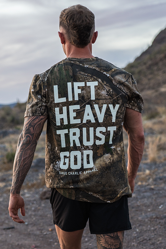 A tattooed man walks outdoors on a rocky path, wearing the Tango Charlie Apparel "Lift Heavy. Trust God." Realtree Premium Camo Tee and black shorts, with mountains and dry grass in the background.