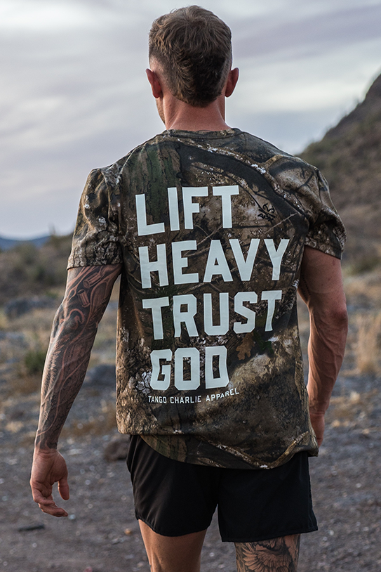A man wearing the Tango Charlie Apparel "Lift Heavy. Trust God." Realtree Premium Camo Tee stands outdoors on a rocky path with his back to the camera, showcasing bold faith-inspired gym wear.