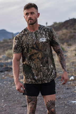 A tattooed man stands outdoors, wearing a Lift Heavy. Trust God. - Realtree Premium Camo Tee by Tango Charlie Apparel and black shorts, with rocky hills and dry vegetation in the background.