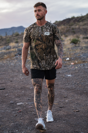 A tattooed man walks on a rocky trail, wearing a Lift Heavy. Trust God. - Realtree Premium Camo Tee by Tango Charlie Apparel, black shorts, and white sneakers. Desert plants and hills are in the background.