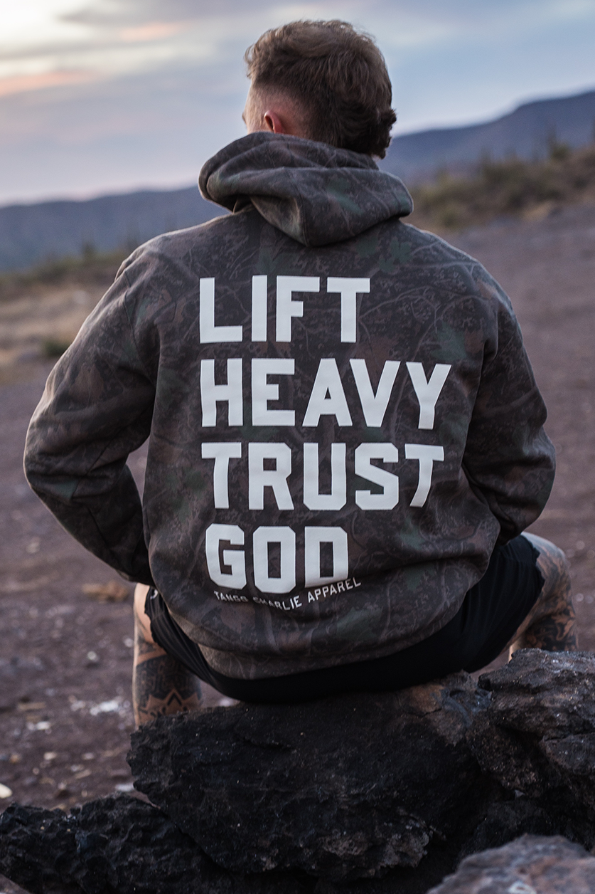 A person sits on a rock in the desert, facing away and wearing a Tango Charlie Apparel “Lift Heavy. Trust God.” Premium Darkwood Camo Hoodie, featuring bold text on the back. With mountains rising in the serene landscape behind them, this Christian fitness enthusiast draws inspiration from Proverbs 3:5-6.