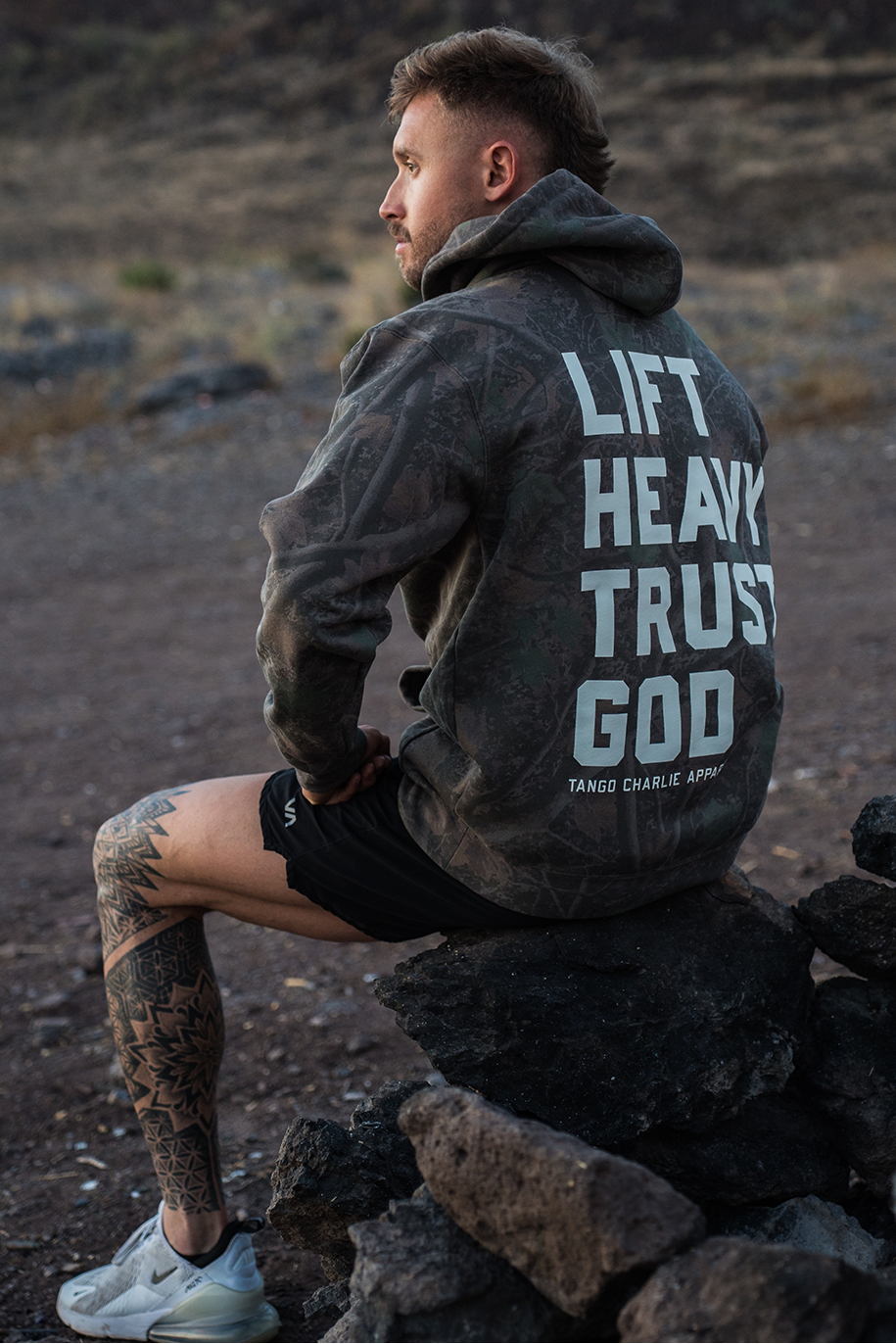 Wearing the Tango Charlie Apparel “Lift Heavy. Trust God.” Premium Darkwood Camo Hoodie, a man sits on rocks outdoors, gazing into the distance and embodying the spirit of Christian fitness inspired by Proverbs 3:5-6.