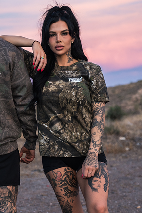 A woman with long dark hair and tattoos wears a Tango Charlie Apparel "Lift Heavy. Trust God." Realtree Premium Camo Tee and black shorts, standing outdoors at sunset with mountains and a pink sky. Another person is partially visible beside her.