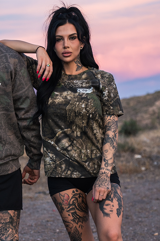 A woman with long dark hair and tattoos wears a Tango Charlie Apparel "Lift Heavy. Trust God." Realtree Premium Camo Tee and black shorts, standing outdoors at sunset with mountains and a pink sky. Another person is partially visible beside her.