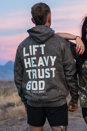 Two people stand outdoors at sunset, one wearing the Tango Charlie Apparel “Lift Heavy. Trust God.” Premium Darkwood Camo Hoodie—capturing the essence of Christian Fitness and inspired by Proverbs 3:5-6.
