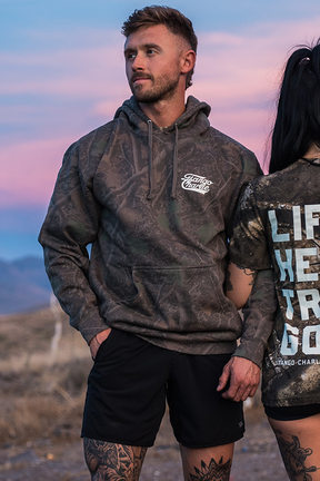 A man in the Lift Heavy. Trust God. - Premium Darkwood Camo Hoodie by Tango Charlie Apparel and black shorts stands outdoors at dusk next to a woman in a graphic t-shirt, both embodying the spirit of Christian Fitness and drawing inspiration from Proverbs 3:5-6.