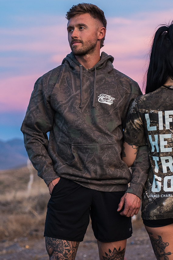 A man in the Lift Heavy. Trust God. - Premium Darkwood Camo Hoodie by Tango Charlie Apparel and black shorts stands outdoors at dusk next to a woman in a graphic t-shirt, both embodying the spirit of Christian Fitness and drawing inspiration from Proverbs 3:5-6.