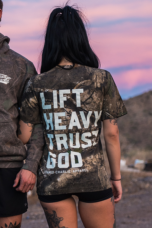 A woman with long black hair stands outdoors at sunset beside another person, wearing a Tango Charlie Apparel "Lift Heavy. Trust God." Realtree Premium Camo Tee. Hills can be seen in the background.