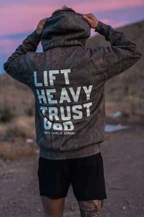 A person stands outdoors with their back to the camera, wearing the Tango Charlie Apparel Lift Heavy. Trust God. - Premium Darkwood Camo Hoodie and black shorts. Against a backdrop of rolling hills and a pink sky, the scene captures the spirit of Christian fitness inspired by Proverbs 3:5-6.