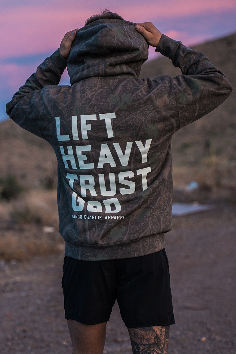 A person stands outdoors with their back to the camera, wearing the Tango Charlie Apparel Lift Heavy. Trust God. - Premium Darkwood Camo Hoodie and black shorts. Against a backdrop of rolling hills and a pink sky, the scene captures the spirit of Christian fitness inspired by Proverbs 3:5-6.