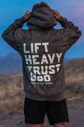 A person stands outdoors at dusk in a rocky desert landscape, facing away while wearing the Lift Heavy. Trust God. - Premium Darkwood Camo Hoodie from Tango Charlie Apparel—a bold statement of Christian fitness inspired by Proverbs 3:5-6.