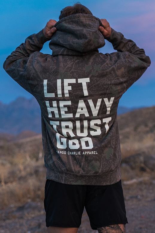 A person stands outdoors at dusk in a rocky desert landscape, facing away while wearing the Lift Heavy. Trust God. - Premium Darkwood Camo Hoodie from Tango Charlie Apparel—a bold statement of Christian fitness inspired by Proverbs 3:5-6.