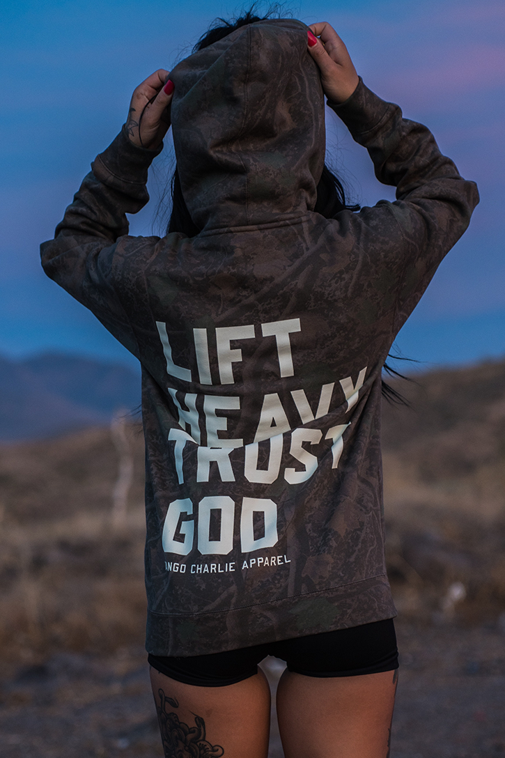 Wearing the Tango Charlie Apparel “Lift Heavy. Trust God. - Premium Darkwood Camo Hoodie,” a person stands outdoors in a natural landscape, holding the hood and embodying Christian fitness inspired by Proverbs 3:5-6. The bold message “LIFT HEAVY TRUST GOD” is printed on the back of this camouflage hoodie.