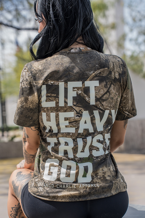 A person with long dark hair sits outdoors in sunlight, wearing a Tango Charlie Apparel "Lift Heavy. Trust God." Realtree Premium Camo Tee, embodying strength and faith.