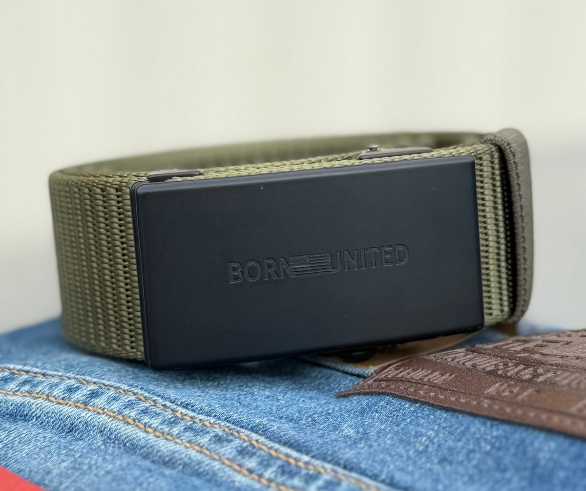 A close-up of the BU Logo Belt - Army Green by Born United Private Label, featuring a black rectangular buckle engraved with BORN UNITED, shown resting on blue jeans with a brown leather patch.