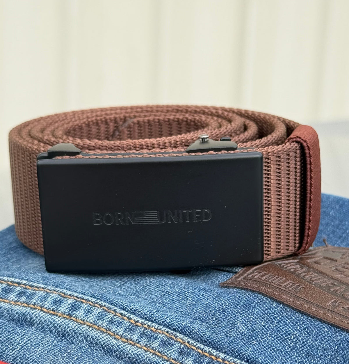 The BU Logo Belt - Brown by Born United Private Label, featuring a black rectangular buckle with the BORN UNITED logo, is shown on blue jeans with a visible Levis leather patch.