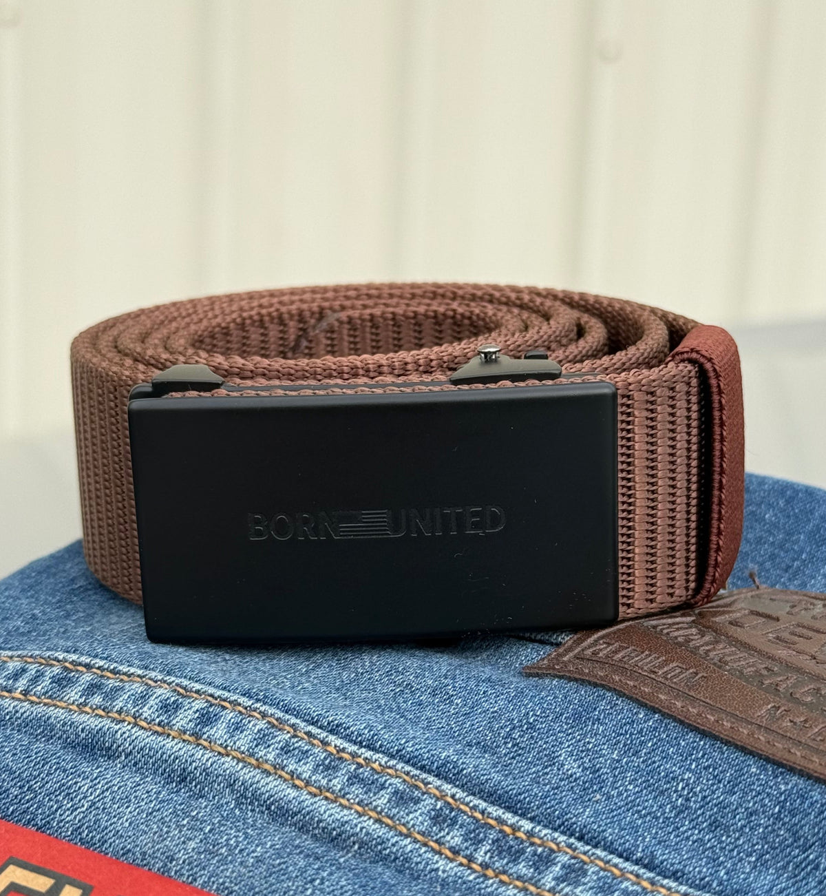 The BU Logo Belt - Brown from Born United Private Label, a brown woven adjustable belt with a black buckle embossed with BORN UNITED, is shown resting on blue jeans with a visible leather patch.
