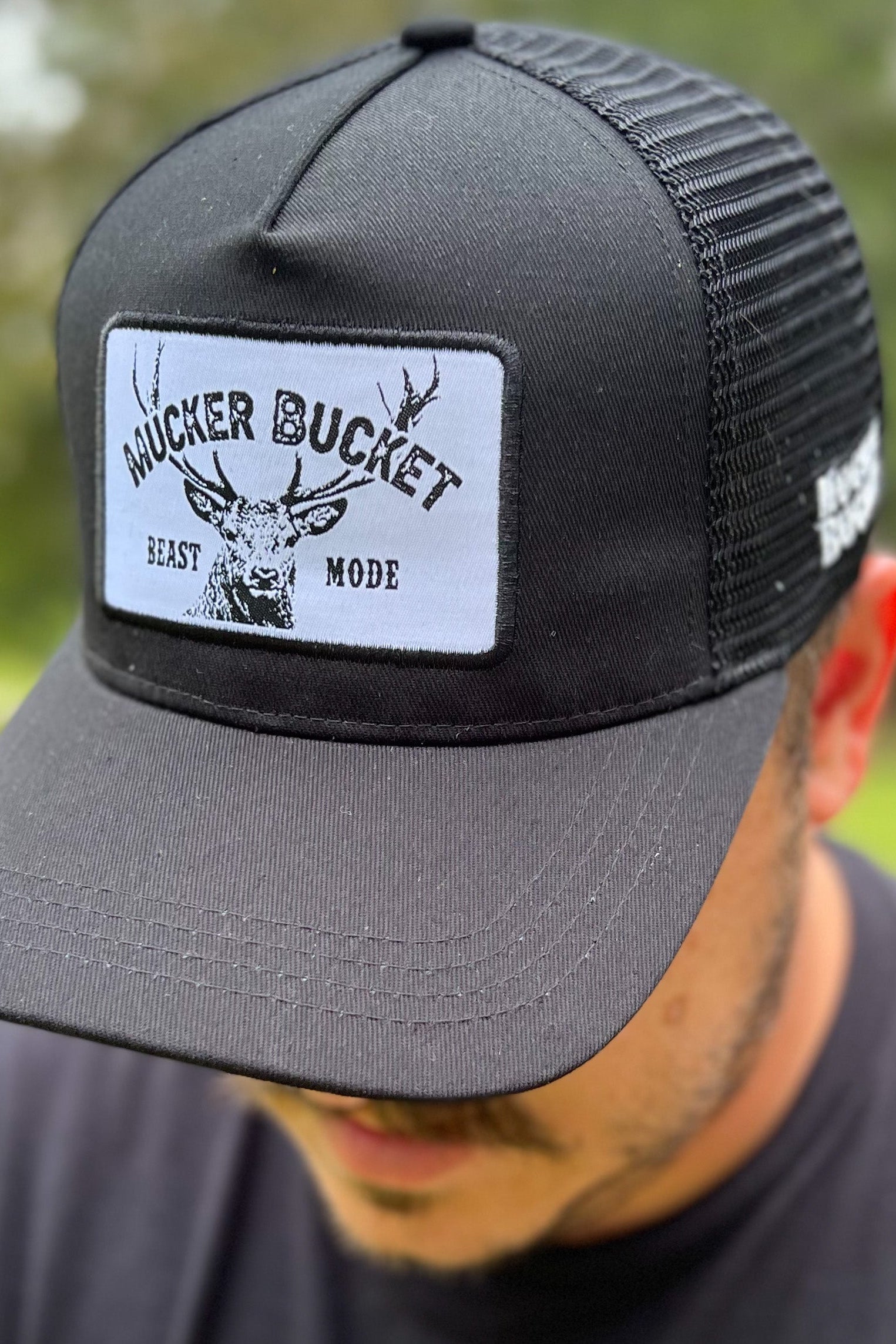 A person wears the Beast Mode OG Trucker Bucket by Mucker Bucket, a black snapback featuring a white-tail deer graphic and patch, against a blurred grassy and tree-filled backdrop.