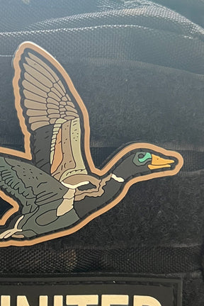 The Mucker Bucket OG Duck It PVC Patch, a colorful 4x4 Velcro patch of a flying duck with outstretched wings, is shown on black fabric partially covering another patch labeled UNITED.