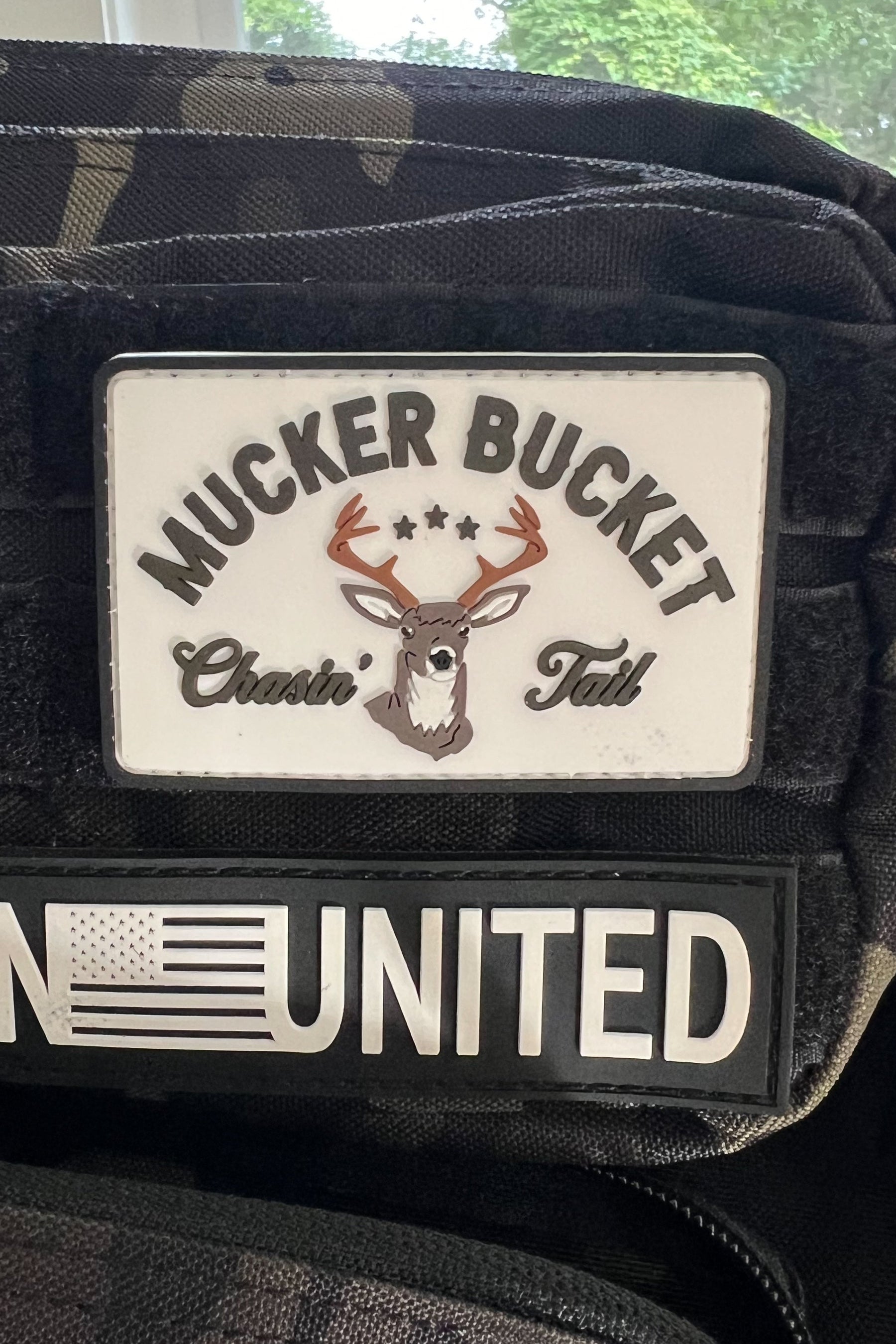 The Mucker Bucket Chasin’ Tail PVC Patch displays three stars, a deer head, and “Chasin’ Tail,” while a 4x4 camouflage patch shows part of the U.S. flag and “UNITED.”.