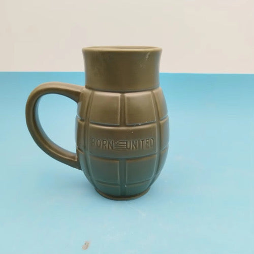 The BU Sip Sip Boom Mug by Born United Private Label is a green ceramic mug shaped like a hand grenade with a handle and “BORN UNITED” embossed on the front, shown on a blue surface against a pale background.