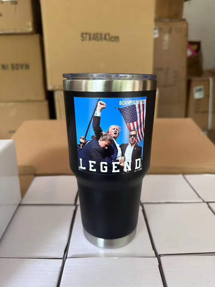The Forever Legend Tumbler - 30oz by Born United Private Label features a black design with a photo of people, one raising a fist holding an American flag, and the words “LEGEND” and “BORN UNITED,” pictured among cardboard boxes.