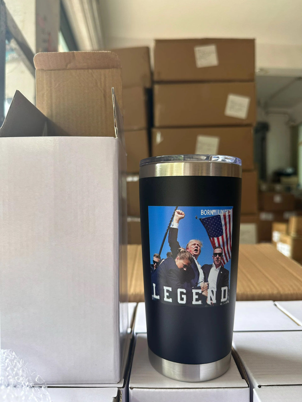 The Born United Private Label Forever Legend Tumbler - 20oz, featuring a photo of a man raising his fist, cheering crowd, and American flag with "LEGEND," stands on stacked boxes in a warehouse filled with more boxes.