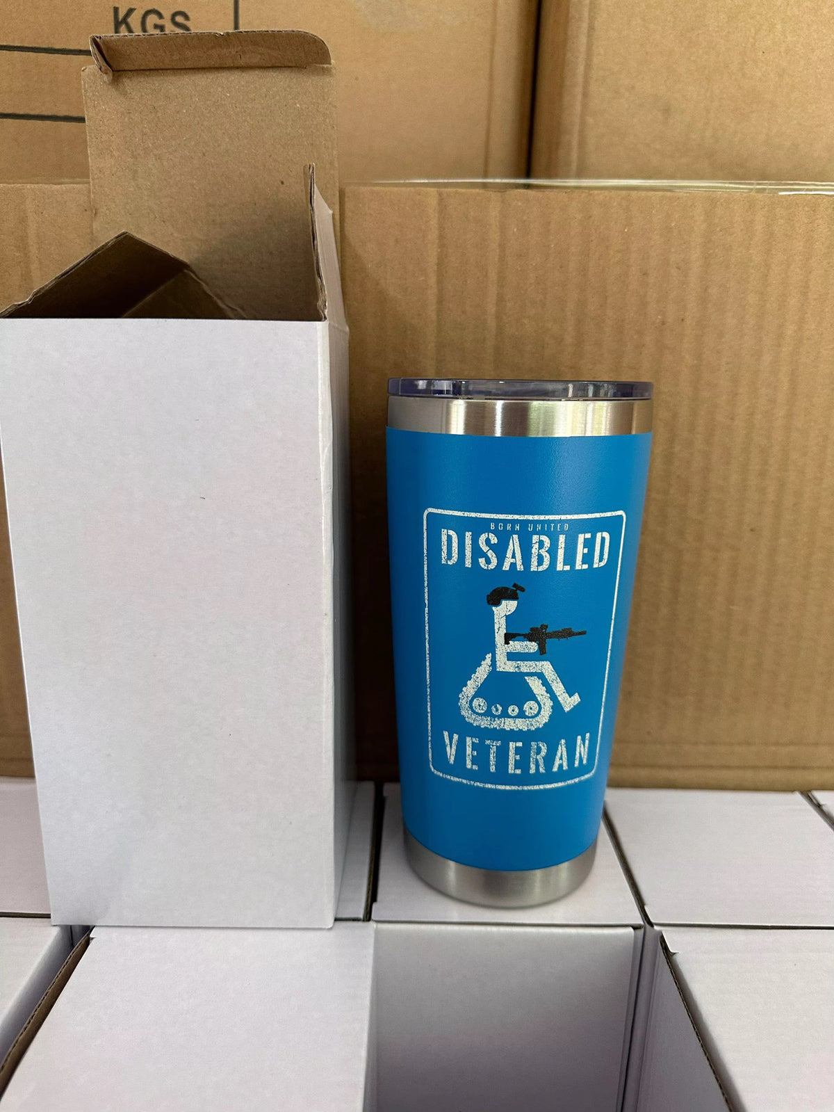 The Born United Private Label Disabled Veteran Tumbler - 20oz, featuring a blue body with a white wheelchair icon holding a rifle and labeled DISABLED VETERAN, is shown beside an open white box on stacked cardboard boxes.