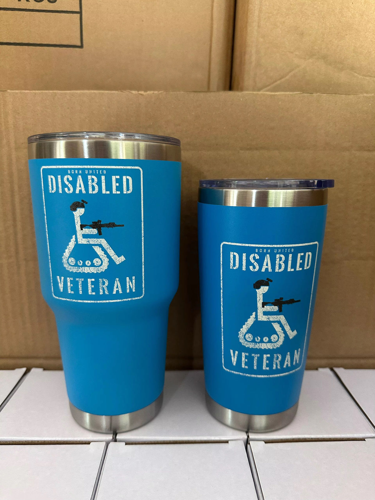 Two blue Born United Private Label Disabled Veteran Tumblers (30oz) with stainless steel bottoms sit side by side, each featuring a white logo of a wheelchair user holding a rifle and the words Disabled Veteran. Cardboard boxes are in the background.