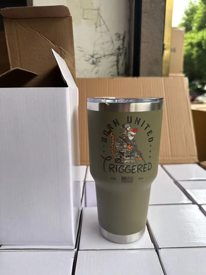 A green 30oz Triggered Tumbler by Born United Private Label, featuring a bear with an American flag and "Born United: Triggered" text, sits on a table near white boxes and an open cardboard box, with trees visible outside.