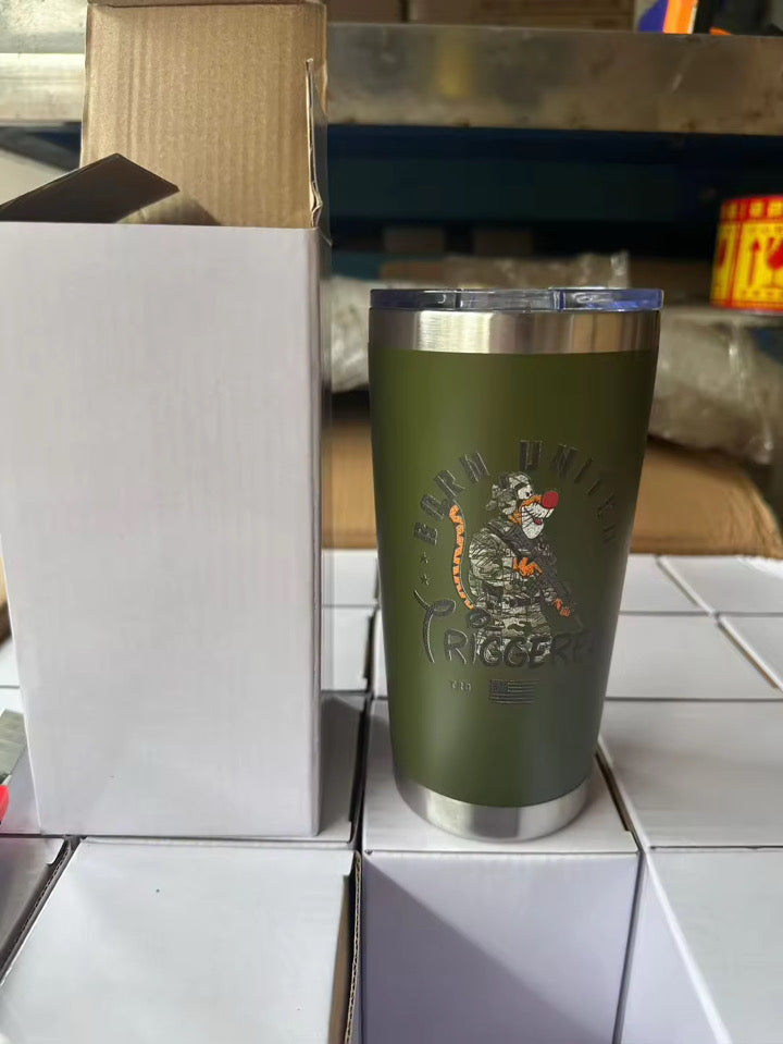 A 20oz Triggered Tumbler by Born United Private Label, featuring an olive green finish with a tiger graphic and Bond Unlimited Triggers text, rests atop white boxes beside a partially opened box in a warehouse.