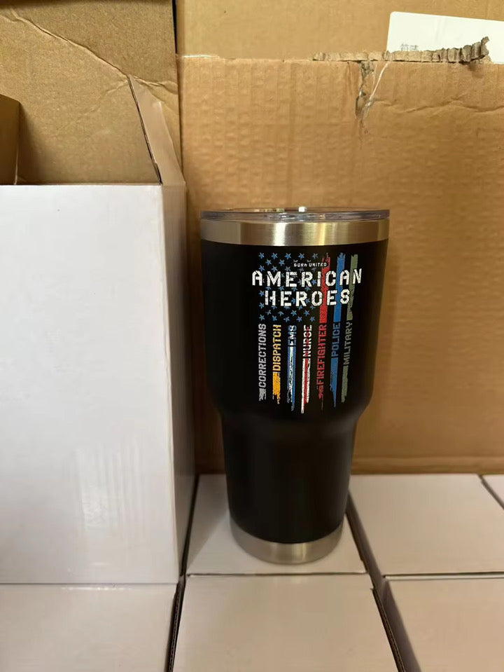 The Born United Private Label American Heroes Tumbler - 30oz features American Heroes, a vertical American flag with colored stripes, and first responder roles printed on a black body, shown in front of cardboard boxes.