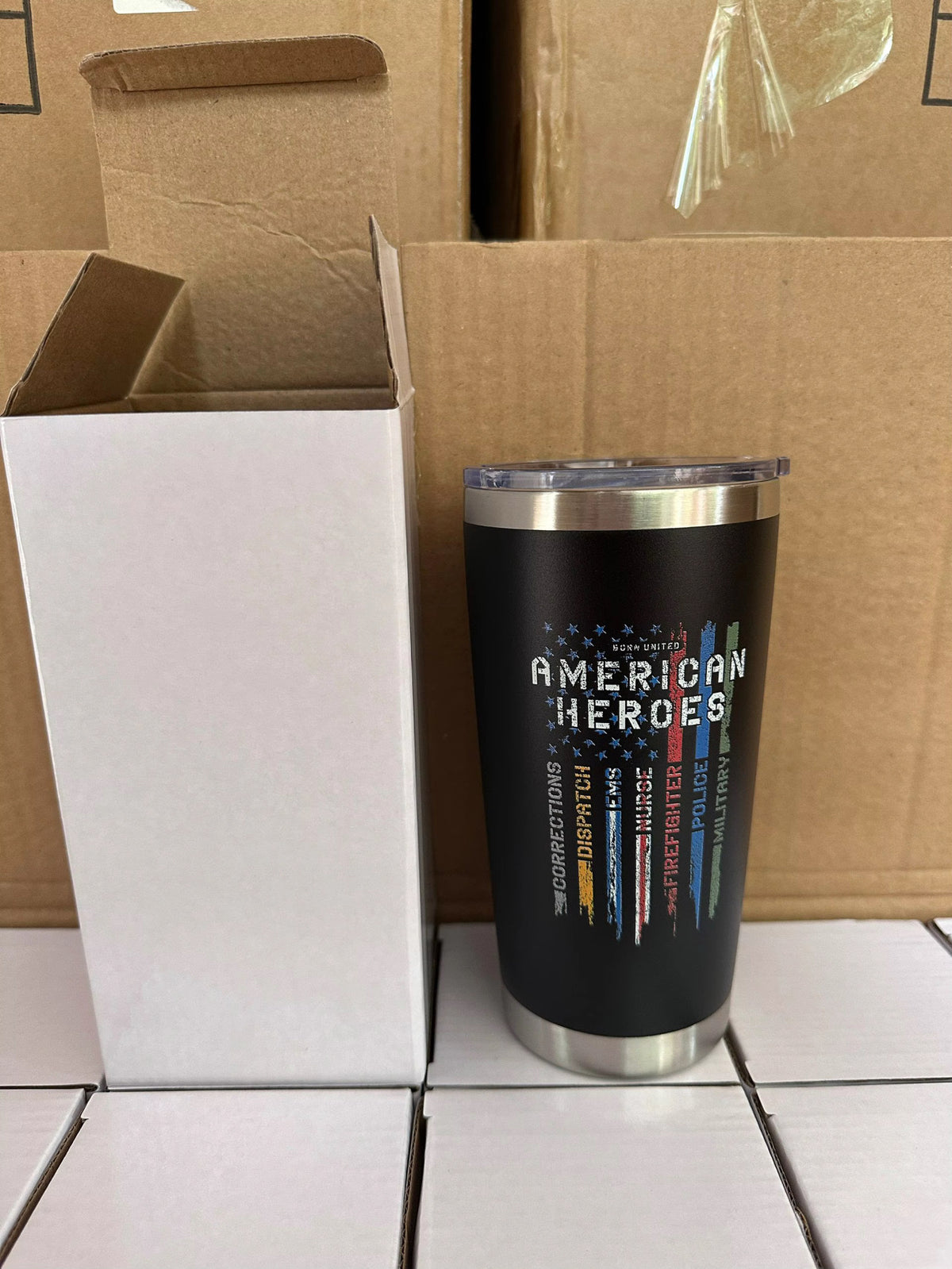 The Born United Private Label American Heroes Tumbler - 20oz, featuring a black design with a distressed flag honoring firefighters, police, and military, stands by an open white box in front of stacked cardboard boxes.