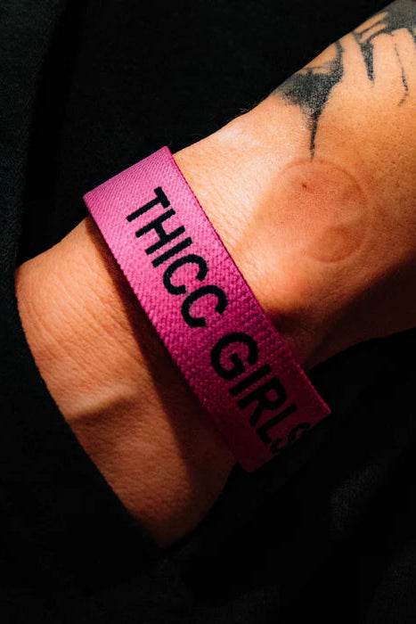 A tattooed wrist showcases the Thicc Girls Club Wristband by Tango Charlie Apparel, featuring bold black THICC GIRLS text on a pink band. Set against a dark backdrop, it celebrates body positivity with every glance.