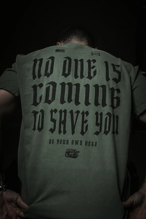 A person faces away from the camera in a dark setting, wearing the Tango Charlie Apparel "No One Is Coming To Save You Tee- Hemp," featuring bold black text: NO ONE IS COMING TO SAVE YOU. GET UP. BE YOUR OWN HERO. Their posture is slightly hunched forward.